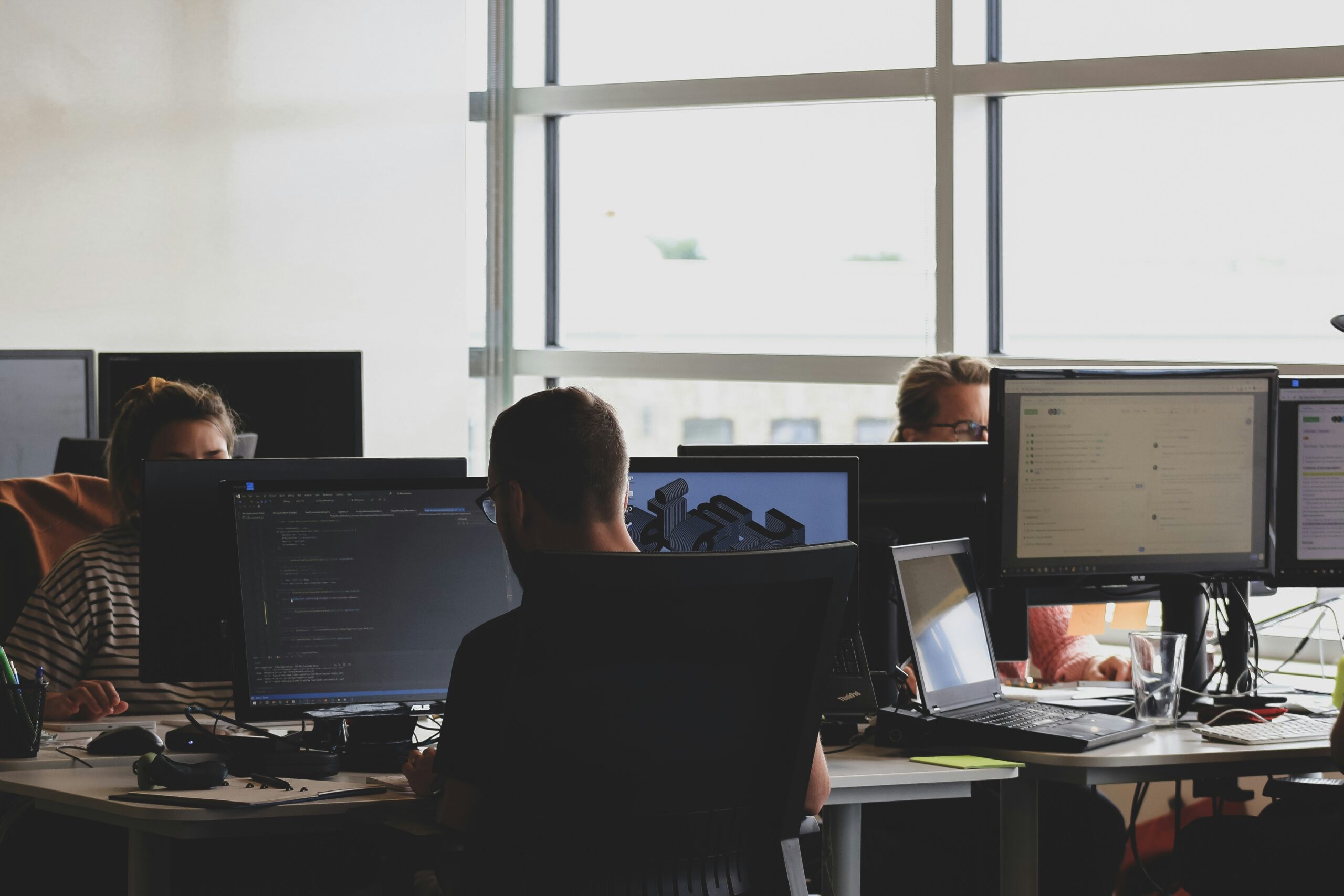 People working at computer stations in a modern office with multiple monitors and natural light.